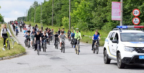 Large group of cyclists riding together on a sunny road, with a police SUV leading on the right.