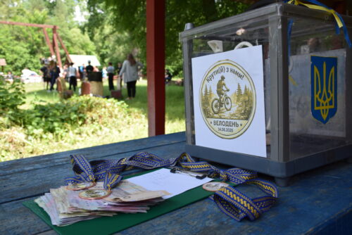 Transparent donation box with Ukrainian insignia on a blue picnic table at an outdoor park event; donation slips and ribbons nearby.