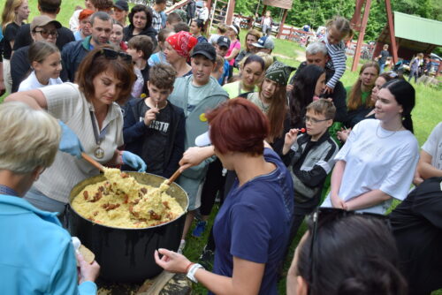 Crowd gathered in a park as a volunteer serves paella from a large pot at an outdoor event.