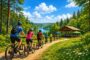 Group of cyclists wearing helmets ride mountain bikes along a dirt trail beside a lake, past a wooden pavilion on a sunny day.