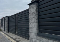 Long black metal fence with concrete stone pillars and a decorative gate along a gravel street nearby a paved sidewalk under a cloudy sky.