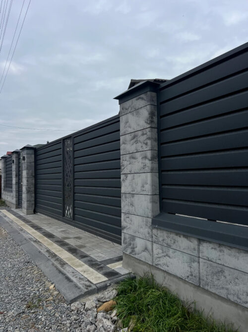Long black metal fence with concrete stone pillars and a decorative gate along a gravel street nearby a paved sidewalk under a cloudy sky.