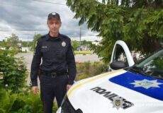 Ukrainian police officer in uniform standing beside a white police car with blue and yellow markings outdoors.