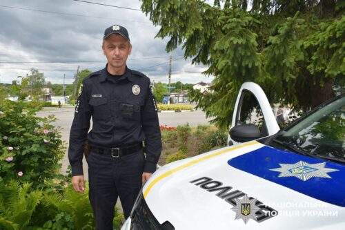 Ukrainian police officer in uniform standing beside a white police car with blue and yellow markings outdoors.