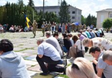 Crowd kneeling in rows during a ceremony while soldiers stand with a Ukrainian flag in a park-like plaza.