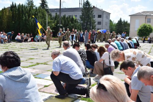 Crowd kneeling in rows during a ceremony while soldiers stand with a Ukrainian flag in a park-like plaza.