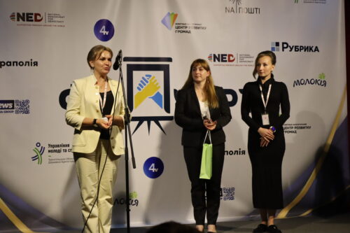 Three women stand on a stage at a conference; one speaks at a microphone while the other two listen, with a logo-filled backdrop behind them.