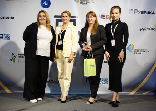 Four women in business attire posing on a stage with sponsor logos behind them; one holds a green gift bag and wears a badge on a lanyard.