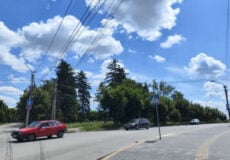 Red car and a dark car at a sunny intersection with a pedestrian crosswalk sign, power lines, and trees against a blue sky with clouds.