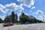 Red car and a dark car at a sunny intersection with a pedestrian crosswalk sign, power lines, and trees against a blue sky with clouds.