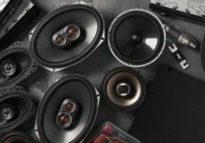 Collection of car audio speakers and components arranged on a dark surface, featuring several large oval woofers, midrange drivers, a tweeter, and wiring.