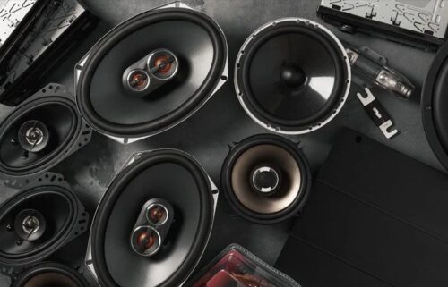 Collection of car audio speakers and components arranged on a dark surface, featuring several large oval woofers, midrange drivers, a tweeter, and wiring.