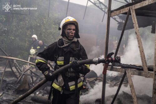 Firefighter in turnout gear hosing down a smoke-filled wooden blaze; another firefighter in the background.