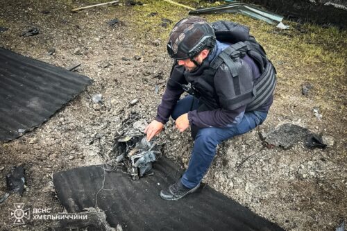 Crouching man wearing a helmet and protective vest inspects a damaged device on rocky ground beside torn black mats.