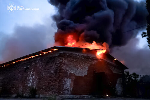 Brick building on fire with flames and thick black smoke pouring from the roof; emergency services logo in the top-left.
