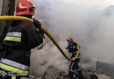 Two firefighters in full turnout gear spray water through a hose amid thick smoke and rubble. Far ground scene of a building fire.