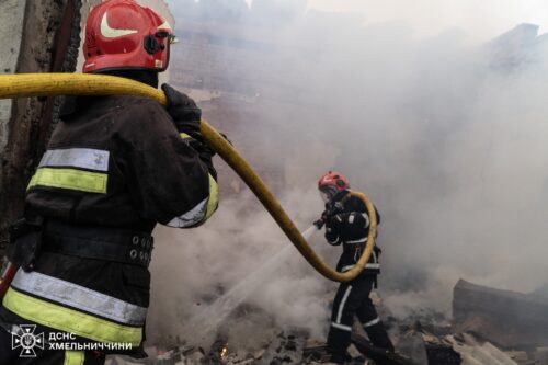 Two firefighters in full turnout gear spray water through a hose amid thick smoke and rubble. Far ground scene of a building fire.