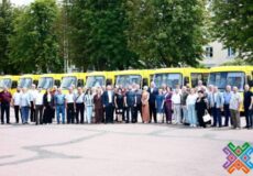 Group of adults standing in a line in front of a row of yellow buses outdoors, with trees behind.