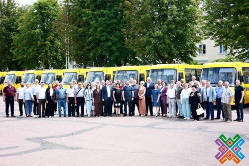 Group of adults standing in a line in front of a row of yellow buses outdoors, with trees behind.