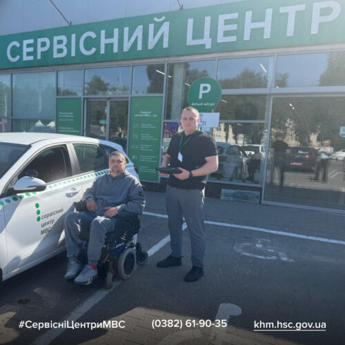Disabled man in a wheelchair and a staff member stand outside a service center with a parked car; large green Ukrainian 'СЕРВІСНИЙ ЦЕНТР' sign visible.