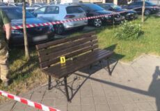 Park bench on a paved area taped off with red-and-white barrier; row of cars behind and a yellow evidence marker labeled '1' on the bench.
