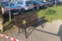 Park bench on a paved area taped off with red-and-white barrier; row of cars behind and a yellow evidence marker labeled '1' on the bench.