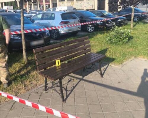 Park bench on a paved area taped off with red-and-white barrier; row of cars behind and a yellow evidence marker labeled '1' on the bench.