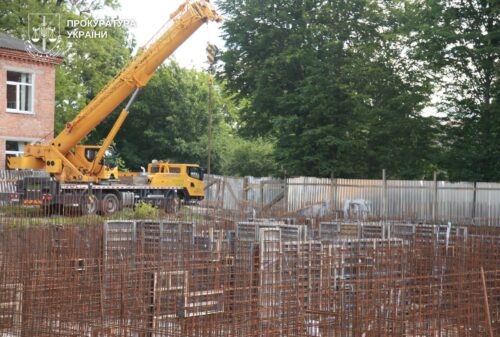 Construction site with a yellow crane truck, rusted rebar cages in the foreground, a wooden fence, and trees in the background; Ukrainian Prosecutor's Office logo in the top-left corner.