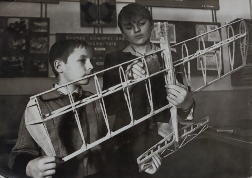 Two boys inspect and assemble a large wooden model airplane wing frame.