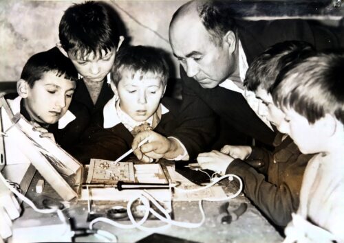 Adults and children gathered around a vintage electronics setup, one man guiding others as they work with wires on a lab bench.