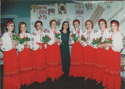 Nine women in Ukrainian folk costumes (white embroidered blouses, red skirts) hold bouquets and pose with a woman in a dark dress in a decorated room.