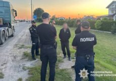 Ukrainian police officers in uniform talk with a man beside a large truck on a roadside at sunset; National Police of Ukraine emblem visible on jacket at right.
