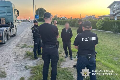 Ukrainian police officers in uniform talk with a man beside a large truck on a roadside at sunset; National Police of Ukraine emblem visible on jacket at right.