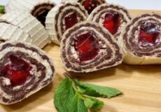 Sliced chocolate Swiss rolls with cream filling and cherry centers on a wooden board, garnished with mint.