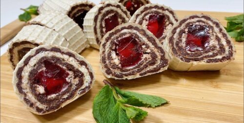 Sliced chocolate Swiss rolls with cream filling and cherry centers on a wooden board, garnished with mint.