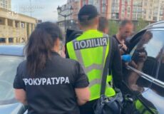 Ukrainian police and prosecutor officers at a vehicle during an operation; backs to camera, jackets read ‘ПОЛІЦІЯ’ and ‘ПРОКУРАТУРА’.