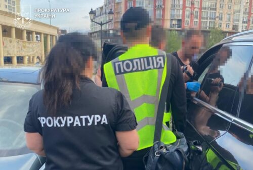 Ukrainian police and prosecutor officers at a vehicle during an operation; backs to camera, jackets read ‘ПОЛІЦІЯ’ and ‘ПРОКУРАТУРА’.