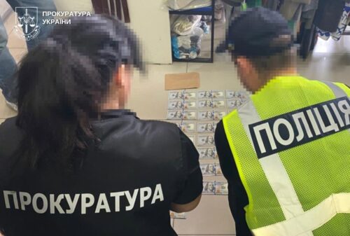Two officers, one in a Prosecutor's Office jacket and one in a police vest, stand with their backs to the camera examining a wall of small ID cards or badges displayed.