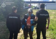 Two Ukrainian law enforcement officers—one from the Prosecutor's Office and one from the Police—restrain a handcuffed man in a grassy outdoor area; faces blurred.