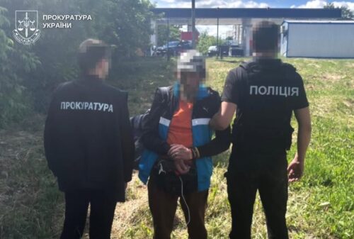 Two Ukrainian law enforcement officers—one from the Prosecutor's Office and one from the Police—restrain a handcuffed man in a grassy outdoor area; faces blurred.
