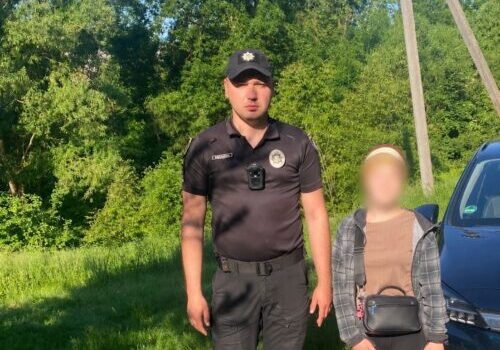 Uniformed officer stands beside a blurred civilian on a gravel path, with a parked car and green trees in bright daylight.