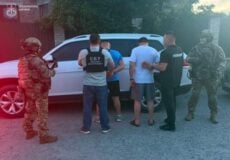 Five men with hands behind their backs are detained by armed officers near a white SUV, with Ukrainian law enforcement logos visible.