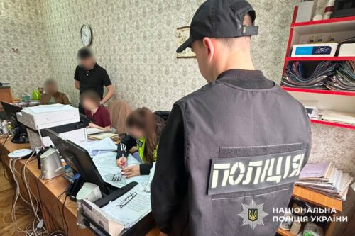 Ukrainian police officer with back to camera in a busy office, seated people at desks filling out forms and coordinating paperwork.