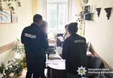 Two Ukrainian police officers in dark uniforms review documents at a cluttered desk in a small office, with plants and a window behind them.