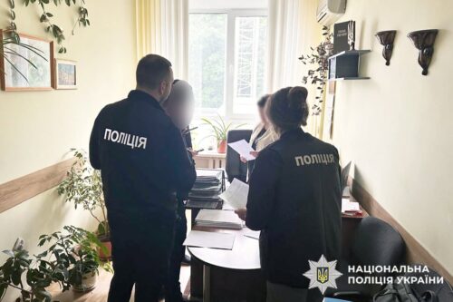 Two Ukrainian police officers in dark uniforms review documents at a cluttered desk in a small office, with plants and a window behind them.