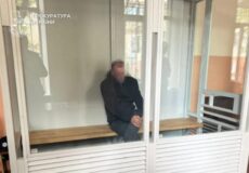 Man sitting on a wooden bench inside a glass-walled detention room at the Prosecutor General's Office of Ukraine (Pochyka/Prosecutor's Office watermark visible).