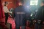 Back view of a man in a dark vest marked with a Ukrainian prosecutor’s office, standing in a press briefing room with cameras and other blurred people nearby.