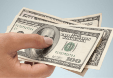 Hand holding several US $100 bills against a pale blue background, fanned out to show the notes