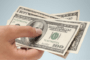 Hand holding several US $100 bills against a pale blue background, fanned out to show the notes