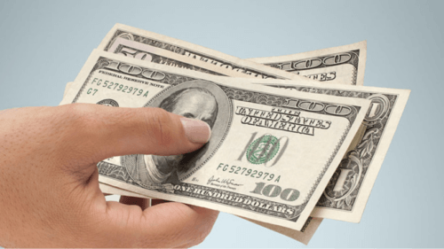 Hand holding several US $100 bills against a pale blue background, fanned out to show the notes